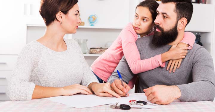 Woman sitting at a table looking at her husband who is signing documents as his daughter hugs him.  Car keys sit on the table