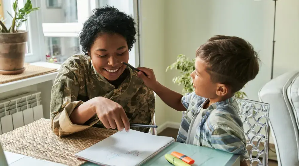 A child seated at a table pretends to paint on the face of a daycare worker as she tries to get him to concentrate on his lesson.