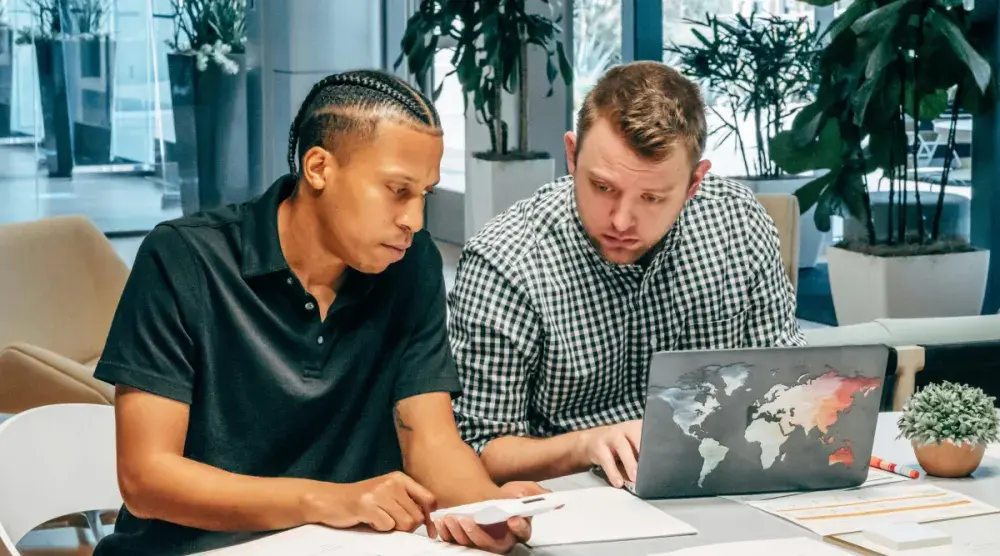 Two young entrepreneurs, one with a calculator and the other with an open laptop computer, sit at a table and calculate the costs of starting an LLC in Illinois.