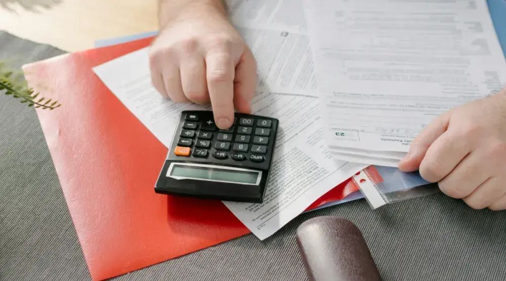 A man sits at his desk with a pile of tax forms and uses a calculator.