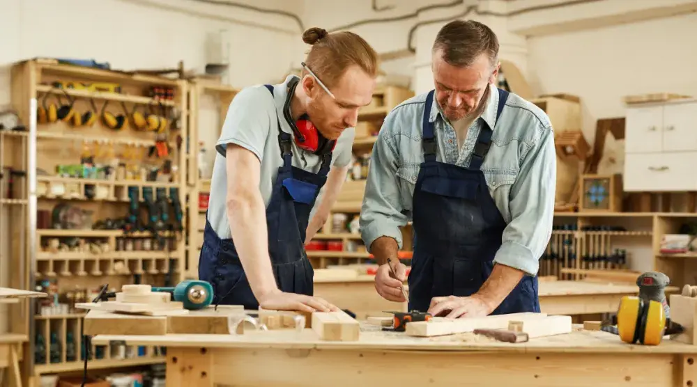 Two carpenters wearing eye and ear protection stand in a wood shop. One shows his slightly younger partner a design.