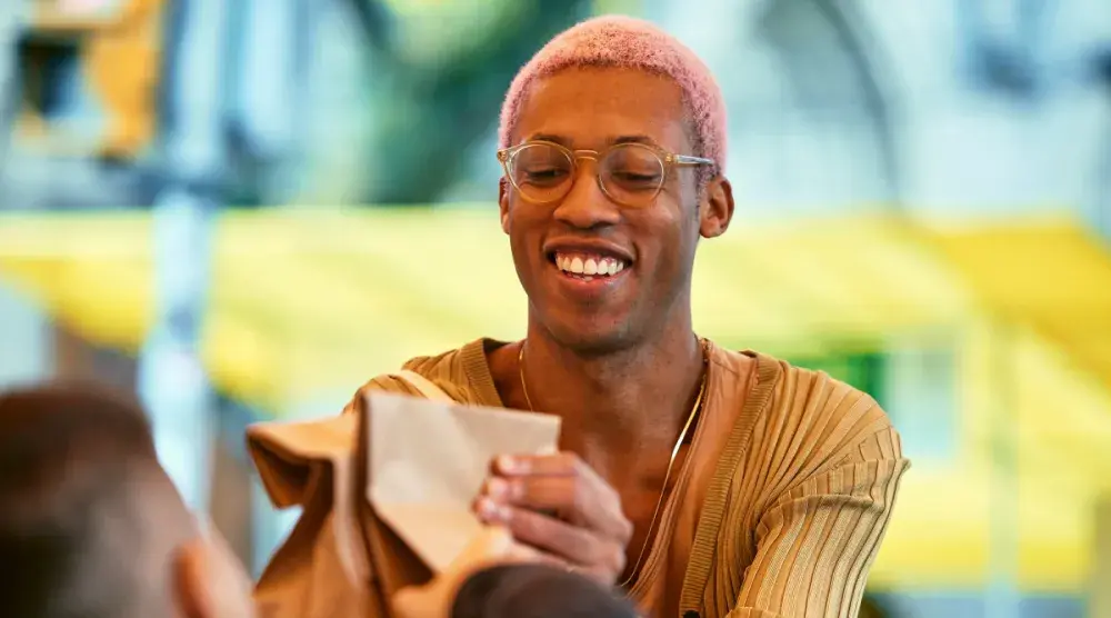 A black man with glasses and pink hair hands a paper bag to his customer at a farmer's market.