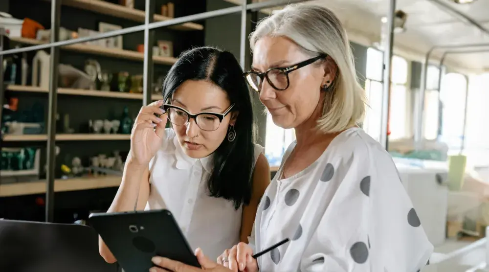 An older white woman shows something to her business partner (a middle-aged Asian woman) on her tablet.