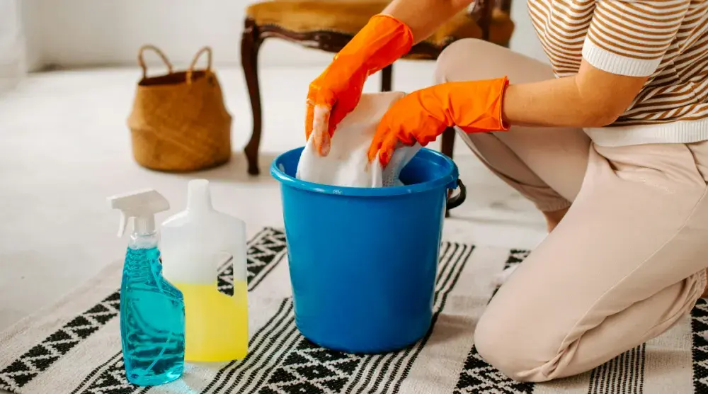 A woman kneels in a modern living room and washes a rag in a bucket next to bottles of cleaning supplies.