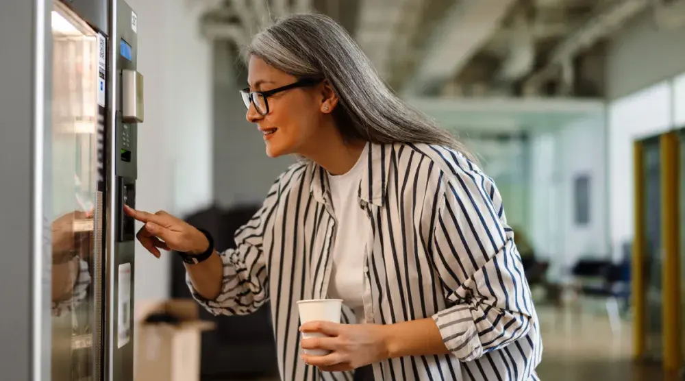 A woman in her 60s holds a coffee cup and presses a button on a vending machine.