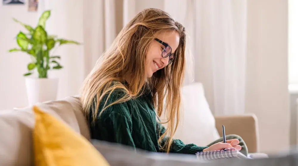 A young woman wearing glasses and seated on a couch smiles as she writes out her last will. In most states, you can be as young as 18 years old to write a legal will.