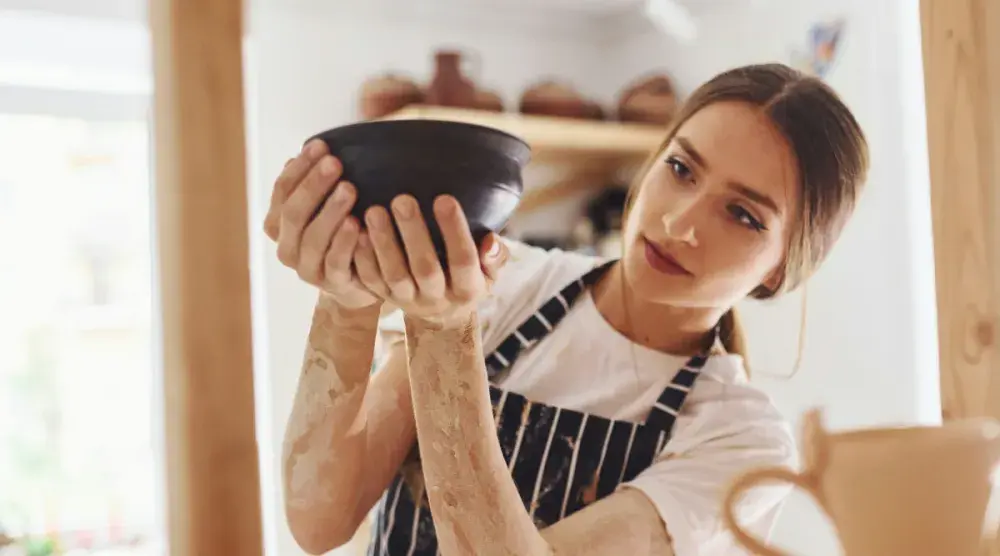 A woman stands in her pottery studio and examines a ceramic bowl.