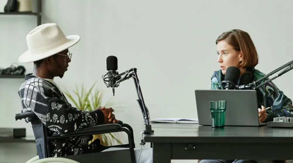 In a bright, professional podcast studio, a woman sits behind a laptop and leans into her podcast mic. She's interviewing a stylish man in a wheelchair.