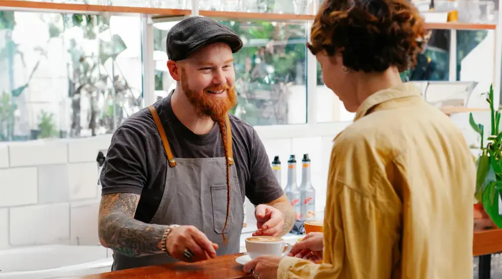 A barista with a long, braided beard hands a cappuccino across the counter to a customer.