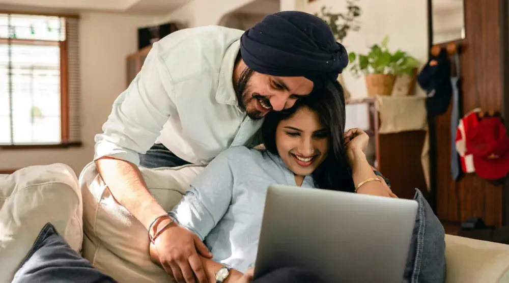 A husband leans over a couch to embrace his wife, who is seated and researching adding a spouse to a house deed on her laptop. Adding your spouse to your deed makes you joint tenants.