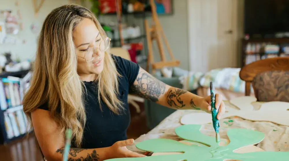 A Hawaiian artist with tattoos sits in her studio and paints a wood carving of a flower.