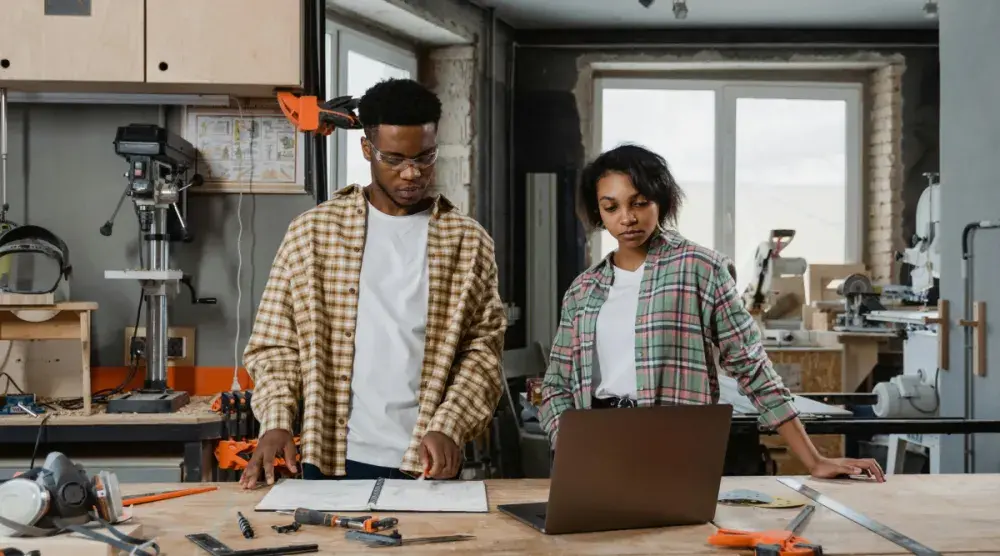 A man and a woman in flannel shirts stand at a work table in their carpentry shop and review LLC formation paperwork on their laptop.