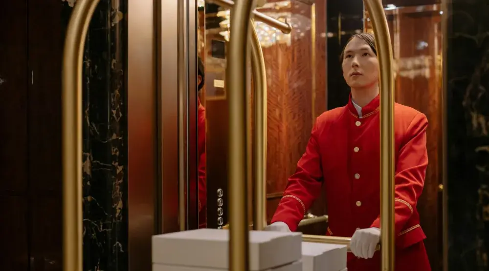 A bellhop in traditional formal dress waits in an elevator with a luggage cart.