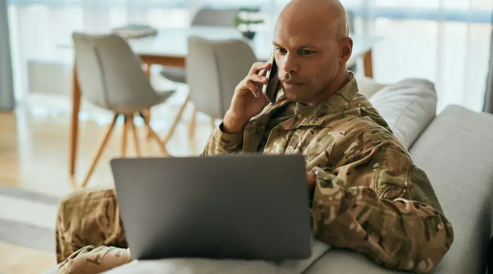 A man in US Military fatigues looks at his laptop and speaks with his lawyer on the phone about his breach of contract case.