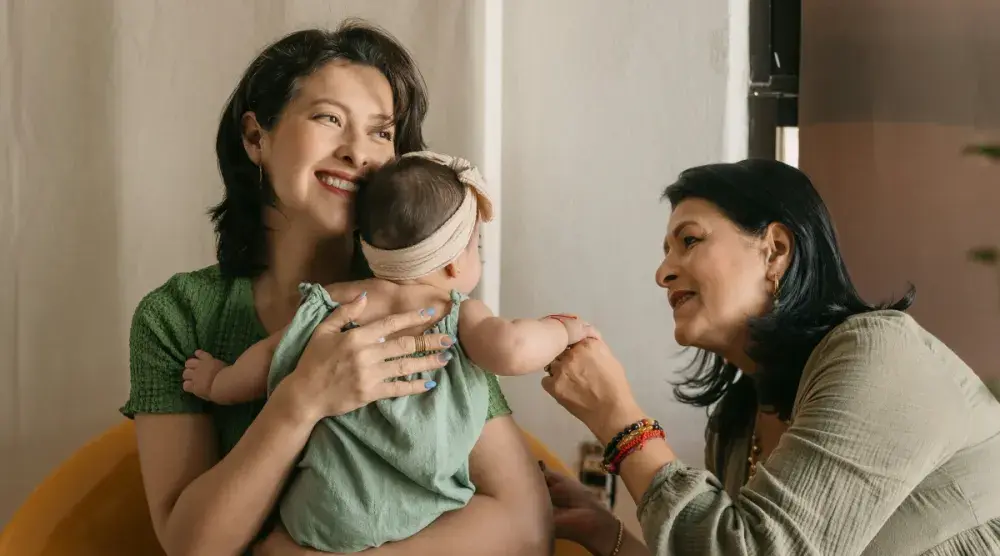 A woman holds her infant daughter while her own mother looks on and plays with the baby's hand.