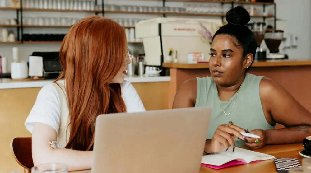 Two women entrepreneurs sit at a table and discuss small business grants to apply for.