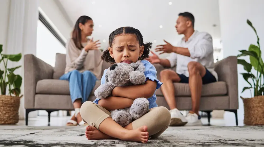 A married man and woman arguing behind a child sitting and clutching a teddy bear. The couple is looking into divorce and alimony options in Pennsylvania.