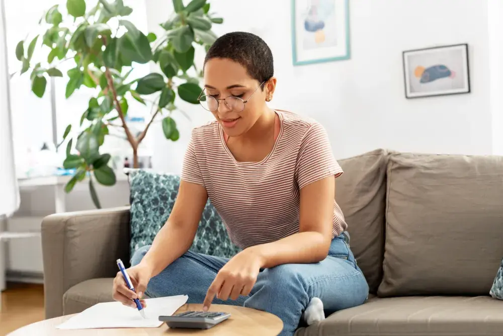 A woman seated on a couch calculates depreciation in a notebook resting on a coffee table.