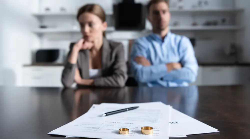 Two gold wedding rings rest on top of divorce papers on a lawyer's conference room table with the ex-spouses seated in the background. Before going through with a divorce, individuals must reflect on whether or not it is their only option.