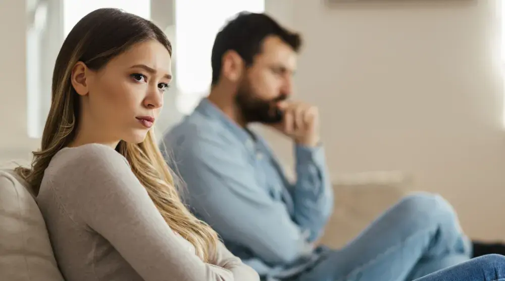 A woman who looks sad sits on one end of a couch in the foreground, while her ex-husband sits at the other end.  The concept of alienation of affection dates back centuries, but remains legal in only six U.S. states.