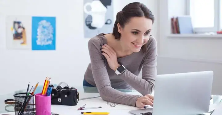 Woman with short brown hair in art studio leans across desk with art supplies and camera on it to type on laptop