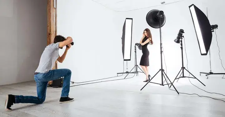 Photographer on his knee taking photos of a woman surrounded by lighting and a white backdrop