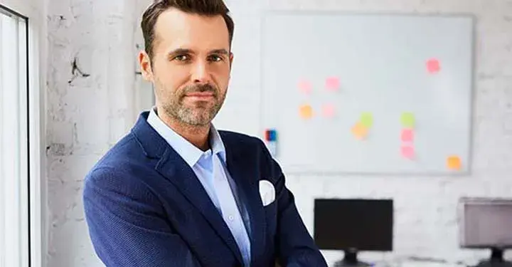 Man standing in suit in office with post-it notes on whiteboard and two computers at desk