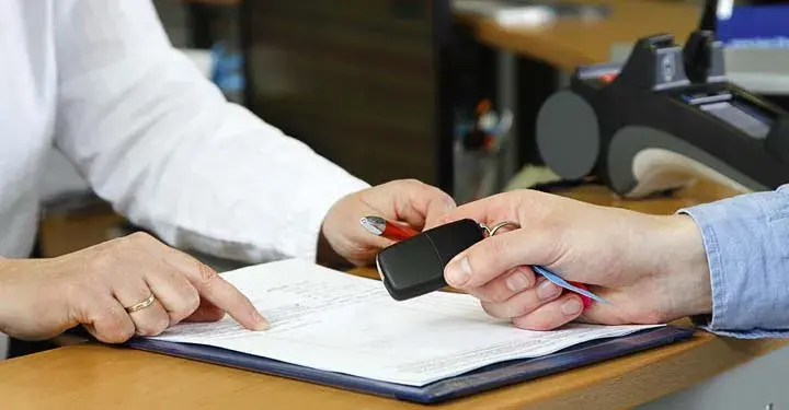 Person's hands pointing at paperwork on a desk.  Another person is holding car keys.