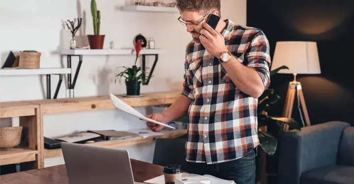 Laid back man in checkered shirt and wire glasses on cellphone in home office looking at document seriously while potted plants sit behind him