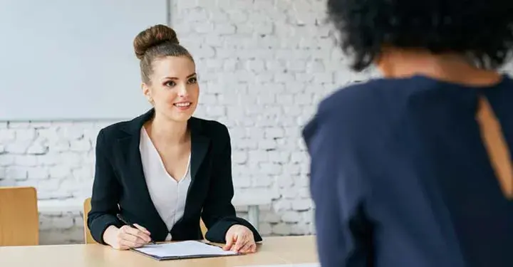 Woman writing on sheet of paper in suit across from other woman