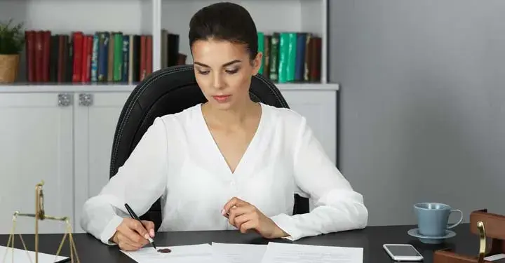 Woman signing forms at desk in front of bookshelf