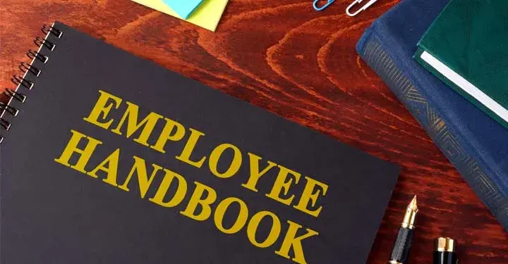 A book whose cover reads "Employee Handbook" sitting on a table surrounded by office supplies