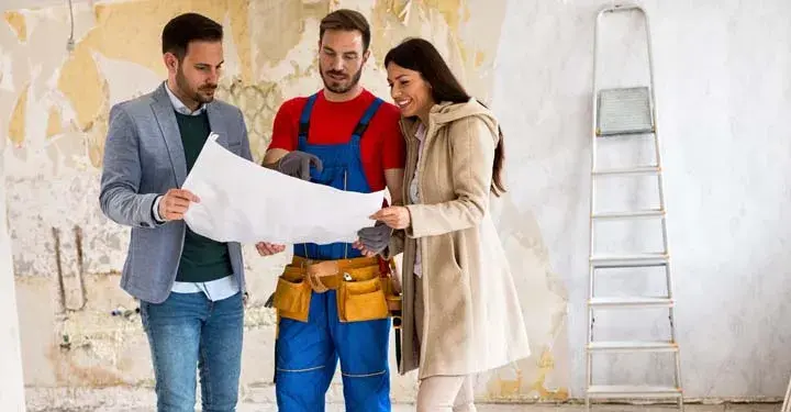 Contractor standing in unfinished house and showing floor plan to couple while wearing tool belt