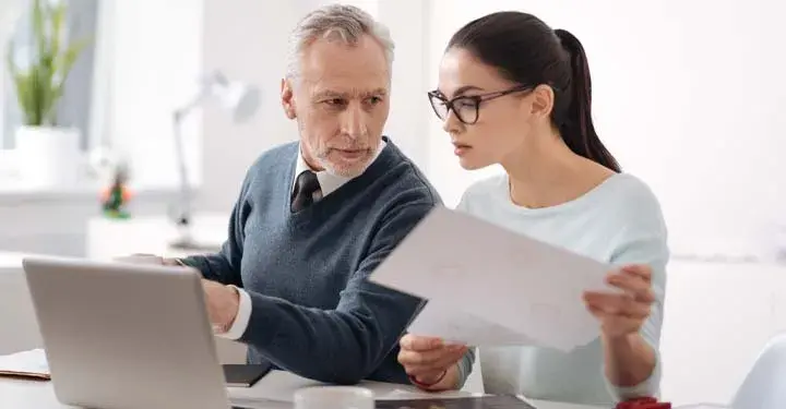 Middle aged man in office points to computer while looking at young woman looking at computer and holding papers