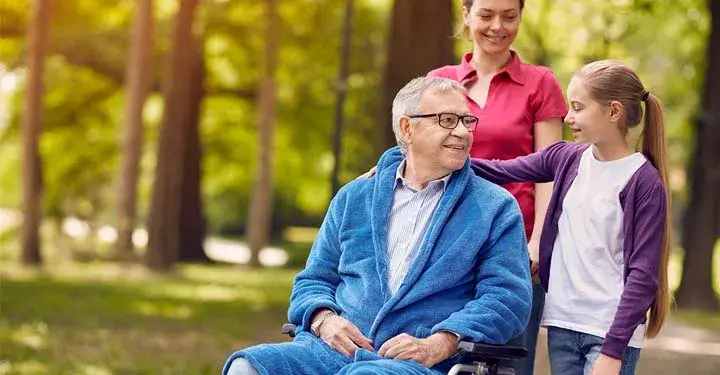 Grandfather and granddaughter smiling at each other in a park as the girl's mother pushes the wheelchair.