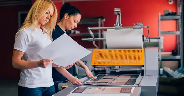 Blonde woman looking at image coming out of machine producing images as second woman stands behind her