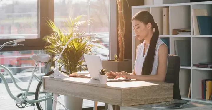 Woman typing on laptop on wooden desk in airy office with powder blue bike resting against full length windows