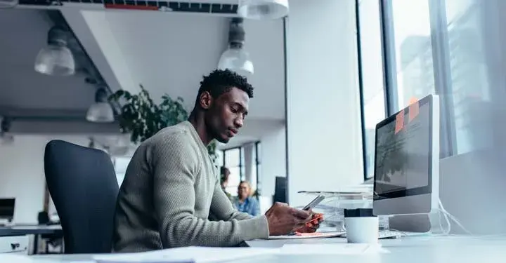 Man looking at smartphone in office in front of desktop computer with coffee mug