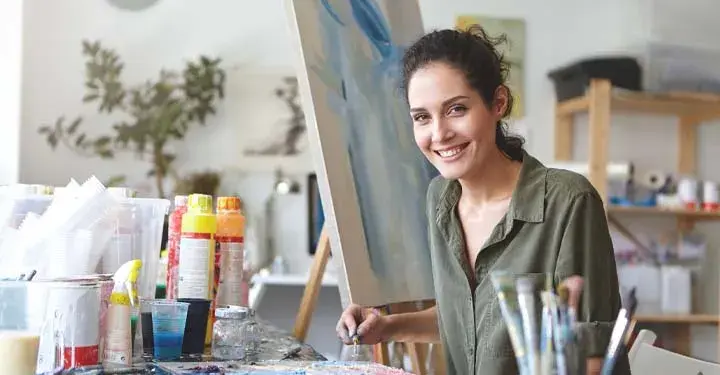 Female artist sitting in studio next to paints and paintbrushes in front of painting on canvas