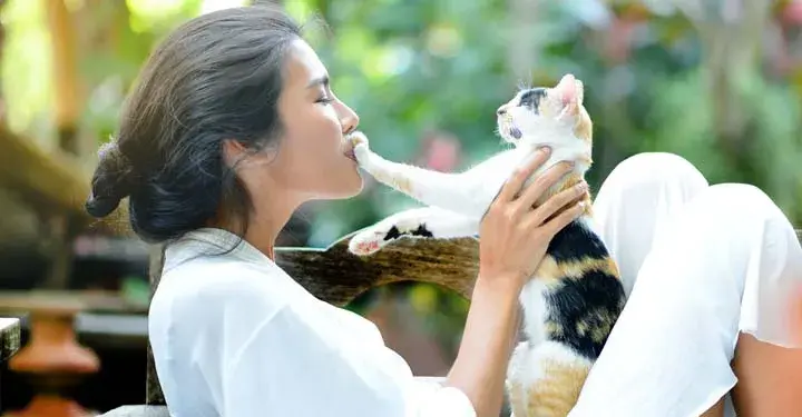 Woman in profile holds up cat and smiles as cat presses paw to her mouth while sitting in park