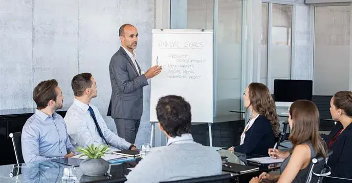 Businessman standing in front of team in conference room gesturing to paper pad that says "annual goals"