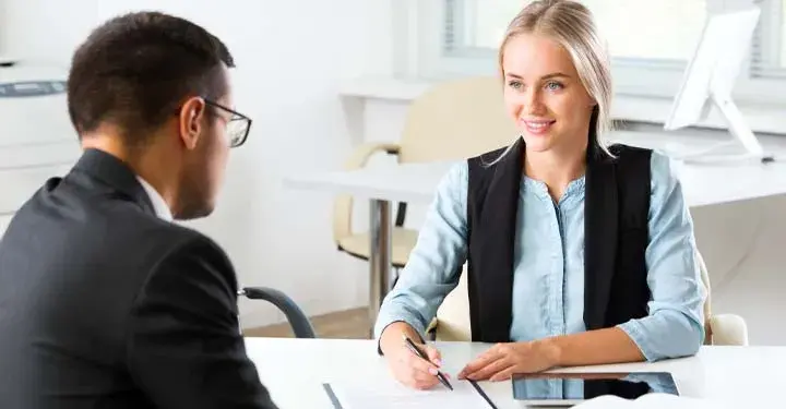 Smiling blonde woman holding pen over document with iPad on desk next <a href=