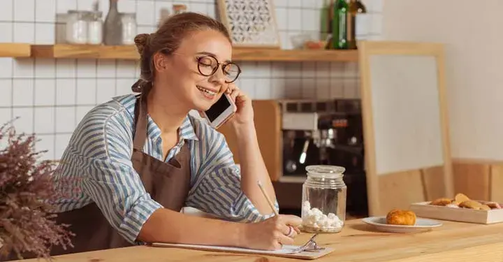 Small business owner sitting in her coffeeshop talking on the phone and writing on a clipboard