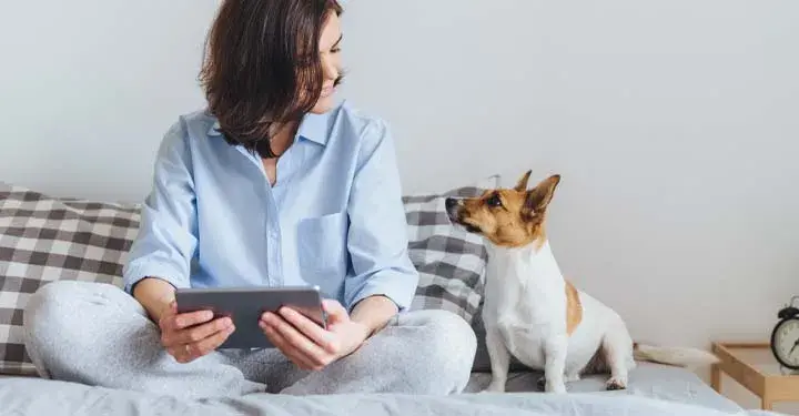 Cross-legged woman on bed looks down fondly at small dog while holding iPad