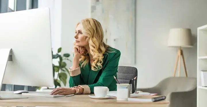 Blonde woman looking at desktop computer with coffee mug and notebook beside it