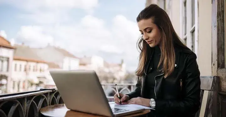 Woman sitting outside on a balcony writing something on a piece of paper next to an open laptop