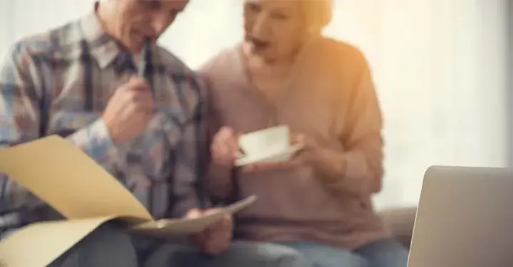 Man holding a pen to his lips next to an elderly woman with a cup of tea reading documents