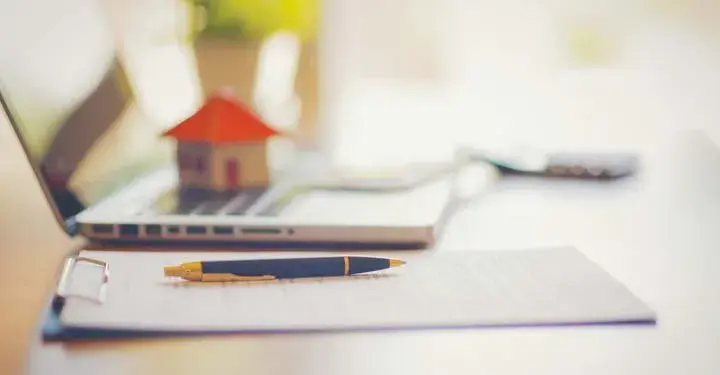 Pen on a clipboard next to a laptop with a small house 