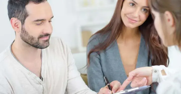 Man signing a document next to a smiling lady while another woman points to the clipboard