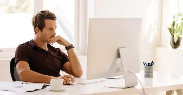 Man with mustache sitting at desk at desktop computer with coffee mug